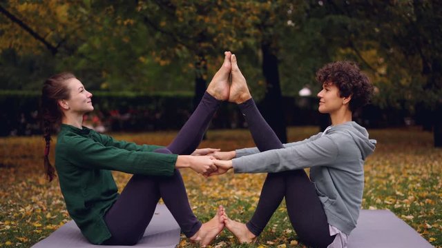 Attractive girls are doing yoga exercises in pair holding hands and putting feet together raising legs sitting on mat in park. Enjoyable training and people concept.