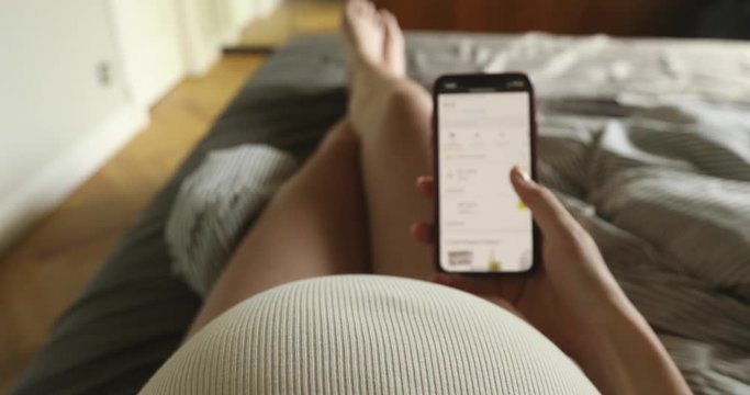 Unrecognizable pregnant woman using smartphone with blank screen sitting on bed. Young expectant lady surfing net, reading about baby care, buying goods for newborn POV
