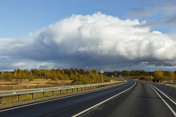 asphalt road among the fields and forests illuminated by the sun, the sky with textured clouds and clouds and a rainbow