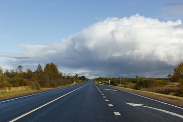 asphalt road among the fields and forests illuminated by the sun, the sky with textured clouds and clouds and a rainbow