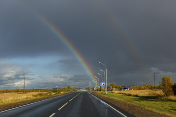 asphalt road among the fields and forests illuminated by the sun, the sky with textured clouds and clouds and a rainbow