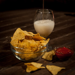 Light beer is poured into a  glass. Next bowl with nachos and sauce sauce with sauce. On a dark wooden table.