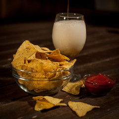 Light beer is poured into a  glass. Next bowl with nachos and sauce sauce with sauce. On a dark wooden table.