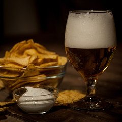 Light beer in a glass on a leg. Next bowl with nachos and sauce  with sauce. On a dark wooden table.