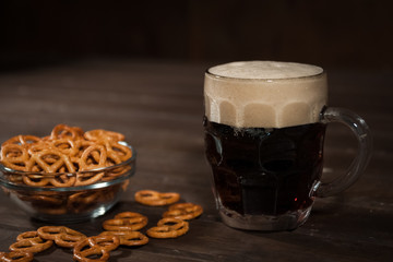 Dark beer in a traditional glass mug. Next bowl with salted pretzels. On a dark wooden table.