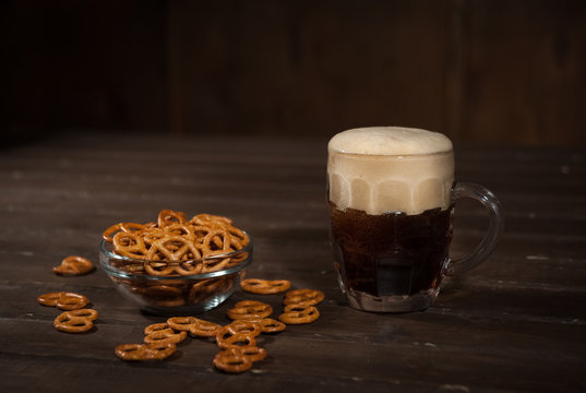 Dark beer in a traditional glass mug. Next bowl with salted pretzels. On a dark wooden table.