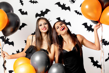 Brunette women in black dresses and witches hats have fun with black and orange balloons on a background with black bats. Halloween