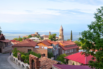 Fototapeta premium Sighnaghi city of love in Georgia, Kakheti region, red roofs of houses and brick towers of the cathedral and fortress, stone road, horizon, blue sky and green trees background