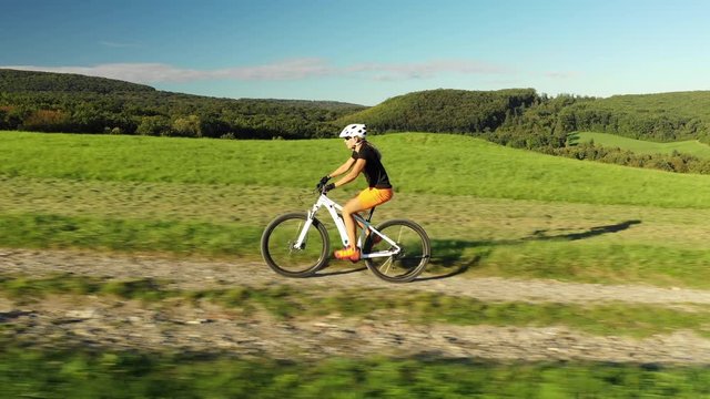 4K sport footage, aerial drone view woman cycling on electric mountain bike up gravel road on hill between meadow
