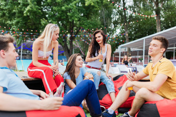 A company of good-looking friends laughing, drinking yellow cocktails and socialising while sitting on the bench and on the lawn in the nice summer cafe. Cheers. Entertainment, having good time