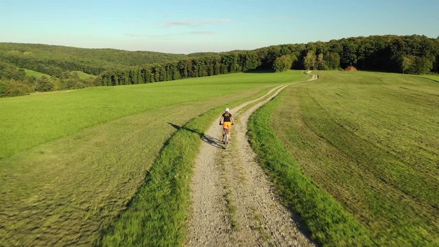 4K sport footage, aerial drone view woman cycling on electric mountain bike down gravel road on hill between meadow
