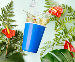 blue cup with tea splashes on tropical background with Red calla Anthurium flowers, fern and monstera leaves. Summer refreshment concept. Text space