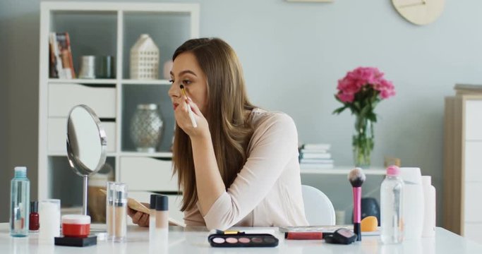 Portrait shot of the pretty young woman doing eyes makeup with shadows and smiling to the camera at the table in her room.