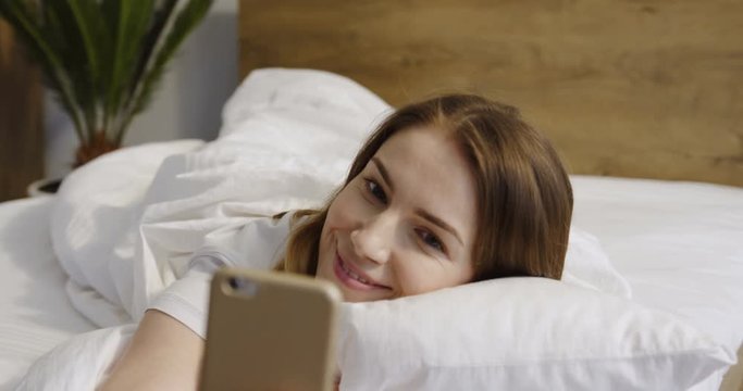 Young Caucasian woman lying in her bed in the morning and taking selphie photos on her smartphone.