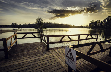 Fototapeta na wymiar Pier with wooden balustrade illuminated by the sunset sun with warning sign to bathe