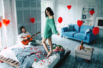 Young cute beautiful woman is dancing on the middle of bedroom while her handsome guy is playing for her on guitar. Love, celebration, romantic, st. Valentine's day, women day, birthday, date. 