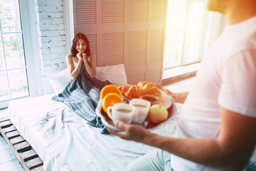 Handsome young man is standing with romantic breakfast on tray, he made it for his beautiful smiling cute girl or wife, who is lying on bed and waiting for him - obrazy, fototapety, plakaty
