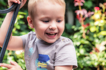 Pretty caucasian laughing baby boy with blue eyes and blonde hair. Summer, outdoor (green leaves and flowers in background).