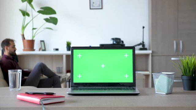 Laptop on the desk with a green screen in the house. A guy is walking in the background and sits on the couch looking at the smartphone