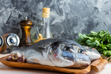 Three are ready to cook raw dorado fish on a wooden tray with herbs, lemon, onions, garlic, spices and olive oil on a gray textured background