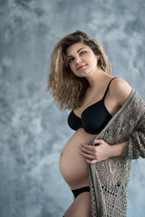 Pregnant woman in black lingerie and knitted cardigan posing on the grey wall background