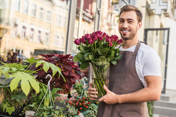 handsome smiling florist holding roses in jar near flower shop