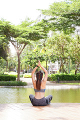 Back view of slim sportswoman holding arms raised and meditating alone on terrace of park pond