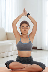 Fototapeta na wymiar Slim Asian woman in sportswear holding hands up and meditating on mat at home