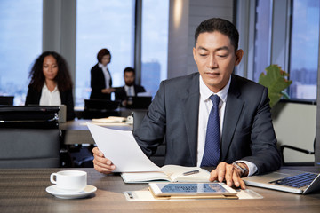 Adult Asian man with tablet and laptop sitting at table and reading papers in office with colleagues