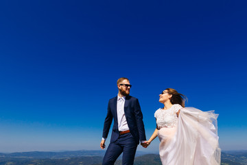 Close-up of newlyweds with sincere emotions who look at each other through sunglasses against the background of the mountains and the blue sky