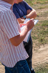 a man in a colored shirt with a notebook in his hands writes with a pen, discusses with another person in the field in the summer agronomist