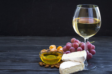 A glass of white wine with cheese cuts, figs, nuts, honey, grapes on a dark rustic wooden boards background