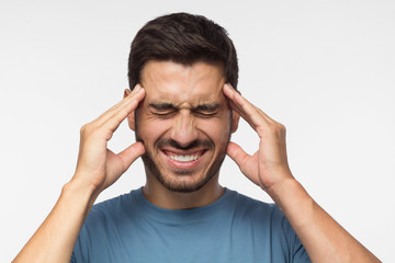Portrait of young man isolated on grey background suffering from severe headache, pressing fingers...