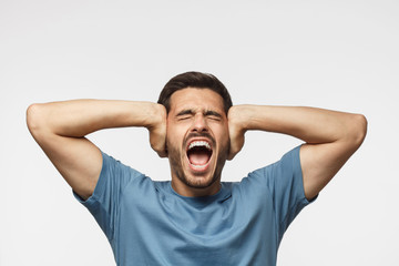 Young handsome man shouting with closed eyes, stressed by noise, closing ears with both hands