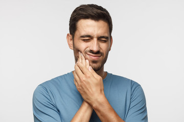 Tooth ache. Young man feeling pain, holding his cheek with both hands, suffering from bad toothache, having painful expression - obrazy, fototapety, plakaty