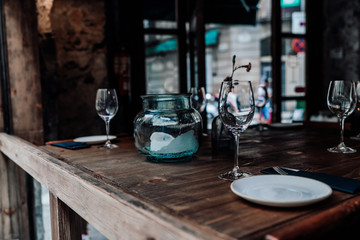 Table setting decor with cutlery, saucers, empty glasses, glass jar with a partially burnt candle inside, wooden table surface; dark interior of a street cafe or cozy restaurant, entrance doors behind