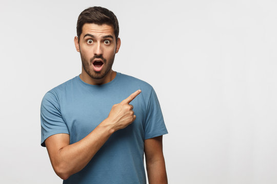 Young surprised man in blue t-shirt pointing right with his finger, shouting WOW, isolated on gray background with copy space fot your text. Shock content