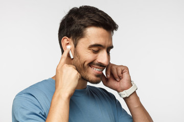 Young man wearing wireless earbuds and blue t shirt, listening to his favorite musical album online, touching one earphone to control application - obrazy, fototapety, plakaty