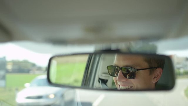SLOW MOTION, CLOSE UP, DOF: Young Caucasian male smiles while driving his car down the road on his way to work. Cool shot of the reflection of a cheerful male driver in the rear view mirror of his car