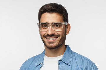 Closeup headshot of smiling young man in blue shirt against gray background, wearing transparent spectacles