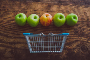 grocery store mini shopping basket with one single red apple among other green ones on wooden table,