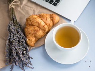 Cup with lavender tea and fresh croissant and laptop on gray background