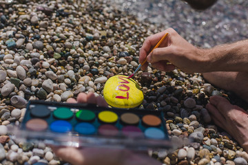 Yellow pebbles on the beach with red inscription love. Love of travel. Valentine's Day. Summer mood. Watercolors are on the beach. Brush and paint. Girl hand draws the word love on pebbles..
