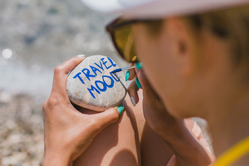 Girl painting on round stone pebble with sign about travel theme. Pebble with sign travel mood. Summer holidays on sea