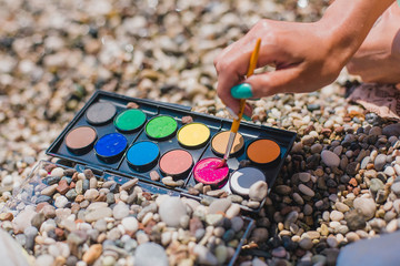 On the beach there is a sea pebble with signs on the theme of travel. Watercolors are on the beach. Drawing on the beach. Artist on vacation.