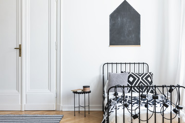 Black poster on white wall above bed in simple bedroom interior with door and table. Real photo