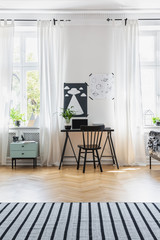 Black chair at desk in bright home office interior with windows, carpet and posters. Real photo
