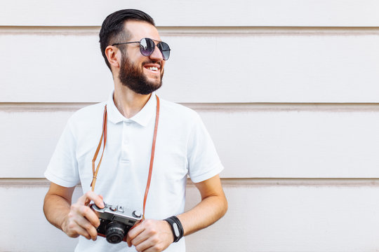 Stylish hipster with a beard and a camera standing near a white wooden wall