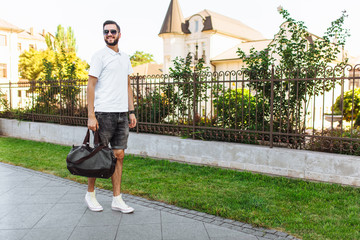 Stylish hipster with a beard in a white t-shirt, with a travel bag in his hands walking around the...