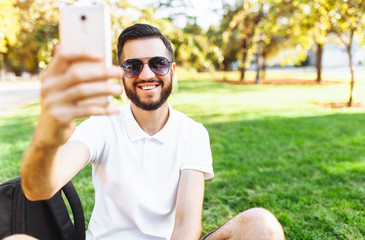 stylish hipster in a white shirt sits on the lawn in the Park and takes a selfie on your smartphone